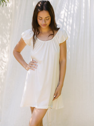 A woman wearing a short-sleeve white nightgown or sleep dress, standing against a white curtain backdrop with soft leaf shadows.