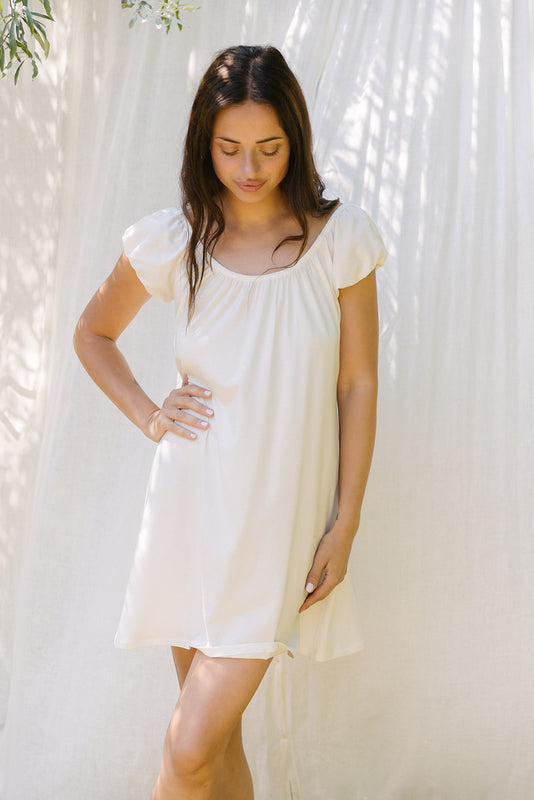 A woman wearing a short-sleeve white nightgown or sleep dress, standing against a white curtain backdrop with soft leaf shadows.