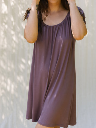 A woman wearing a loose, short-sleeve scoop-neck dress in a muted dusty plum purple, standing against a light wall with soft leaf shadows.