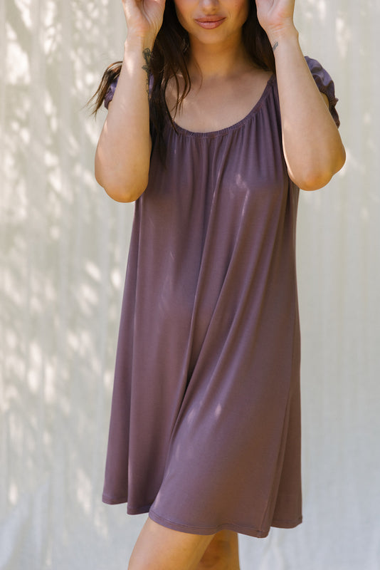 A woman wearing a loose, short-sleeve scoop-neck dress in a muted dusty plum purple, standing against a light wall with soft leaf shadows.